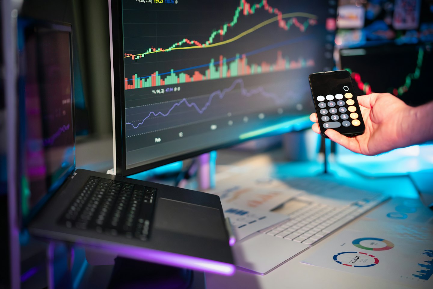 Una mano sostiene un teléfono que muestra una calculadora cerca de datos financieros en monitores.